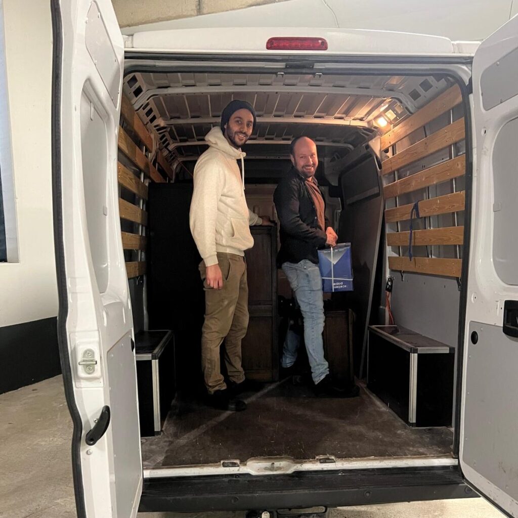 Ammar and David loading an old wooden cabinet and equipment into a white transport van.