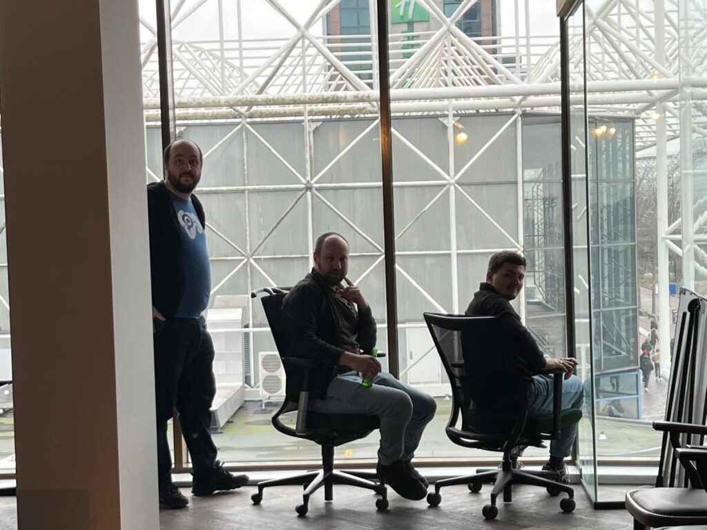 From the left Sorin, David and Alex sitting in office chairs inside a modern building with large windows overlooking a glass and metal structure.
